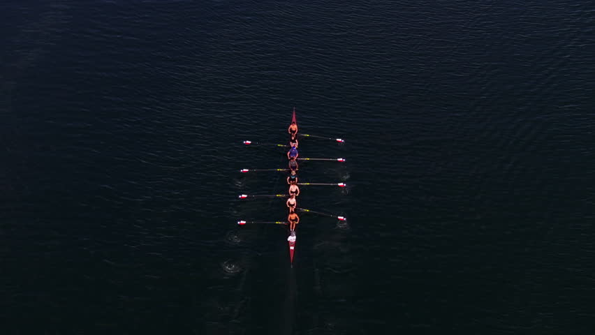Aerial view of rowing team synchronizing strokes on calm waters at twilight, creating ripples on the lake. Sports and outdoor concept