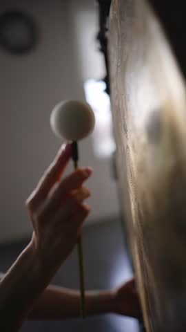 Woman makes the sound of a gong with small soft mallet in yoga studio. Sound healing therapy indoors. 
