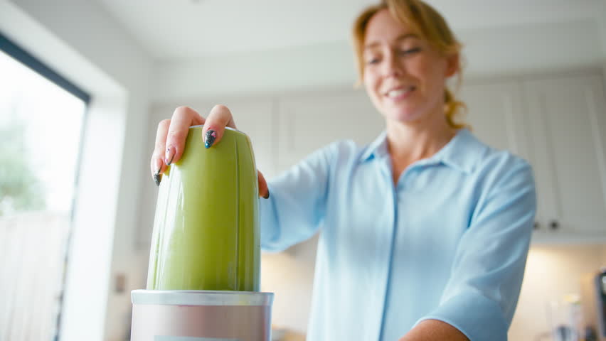 Woman pouring and drinking fresh healthy green smoothie made from apples,banana and celery from blender into glass in kitchen at home - shot in slow motion