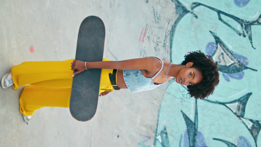 African model posing with skateboard on urban skate park with graffiti. Beautiful curly girl looking camera holding longboard outdoors vertically video. Gorgeous black haired woman enjoy extreme hobby