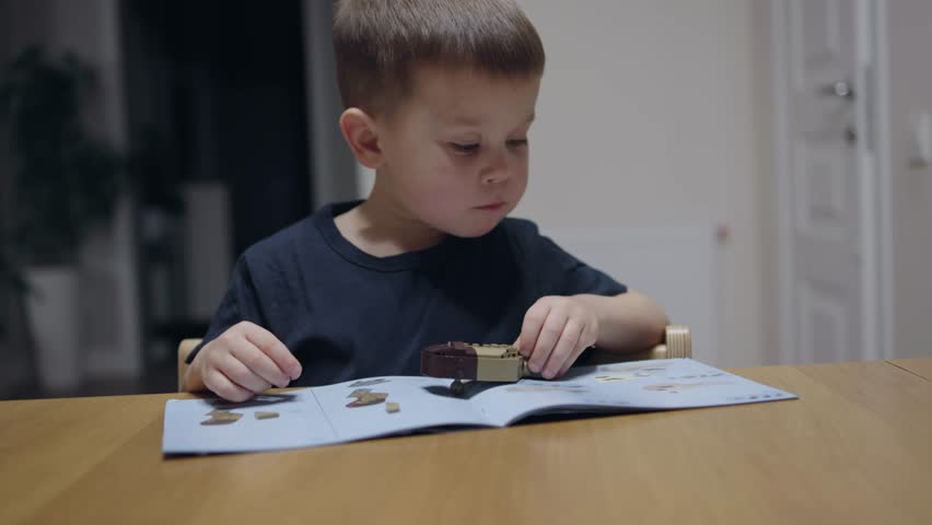 Cute boy sits at table playing educational toy construction set following instructions to assemble bricks. Boy learning world of building blocks in toy construction set experience of toy constructor