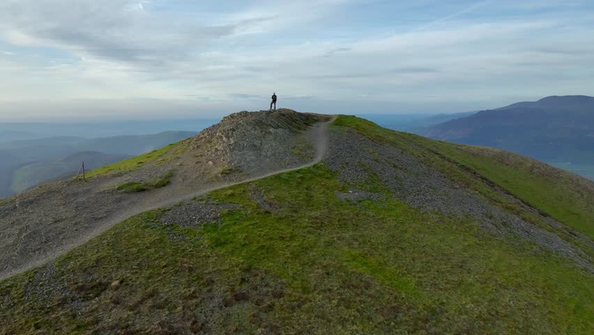 Lone mountain walker at winding path end on fell summit at golden hour. Camera rise revealing forested fells beyond. Grisedale Pike, Lake District, Cumbria, England, UK