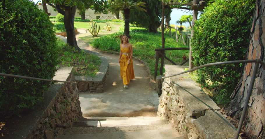 A woman climbs stone steps in a garden, smiling joyfully with lush greenery around her.