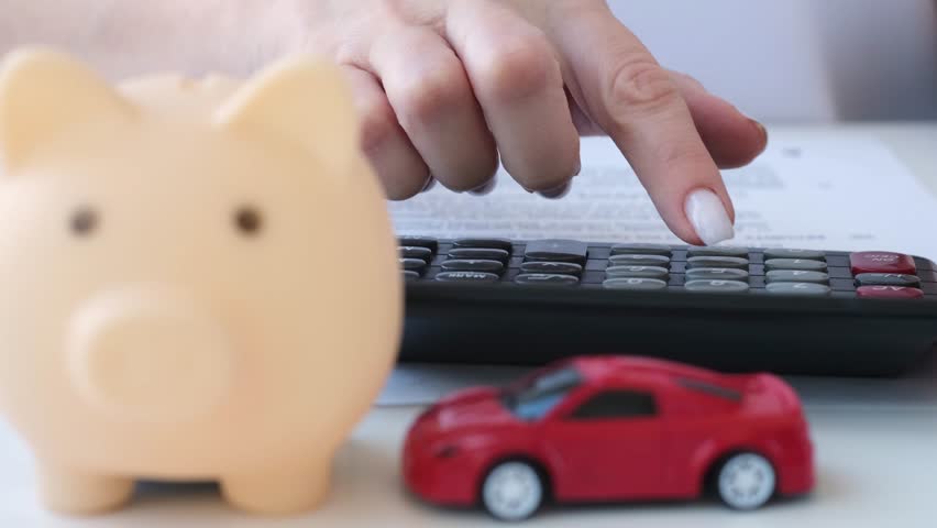 Close up blurred piggy bank and red car, female hand counting on calculator and writing on background. Saving to buy auto, automobile insurance, expenses and tax concept