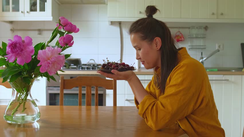 A girl eats cherries sitting in a cozy kitchen, there is a bouquet of peonies on the table. High quality 4k footage