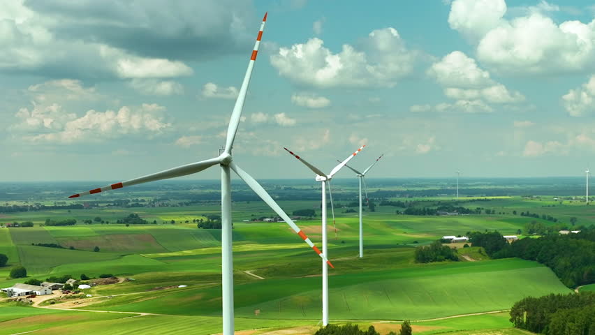 Aerial highlighting a row of wind turbines standing over green agricultural fields. The expansive rural landscape stretches into the distance under a sky filled with fluffy clouds, sustainable energy