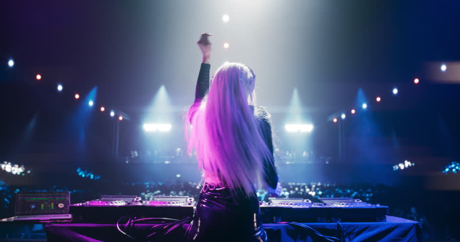Female DJ Delivers a Live Performance of Electronic Music while Standing Behind Mixing Decks at a Massive Event. Colorful Lights and Dance Beats Play at an EDM Concert. Footage from the Back