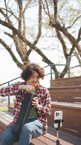 Curly brunette man blogger in casual clothing advertises a tripod in front of the camera outdoors