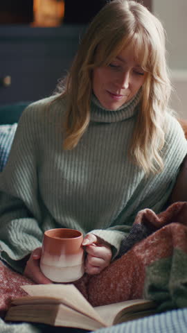 Vertical video close up of woman at home in winter jumper with warming hot drink of coffee in mug reading book - shot in slow motion