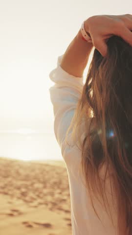 Relaxed woman admiring sunrise on sea coast in summer vacation, touching her long hair, back view. Female traveler enjoying sunset in beautiful beach, vertical video, slow motion, weekend trip to sea