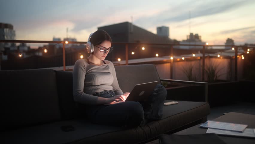 Woman working on laptop outdoors at twilight in a modern city setting, a focused freelancer. A woman with headphones is sitting on a rooftop sofa, working on her laptop as the sunset - Powered by Shutterstock - Get 15% off with code: PIKWIZARD15