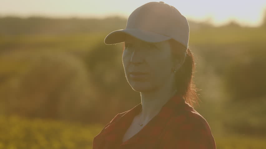Portrait of pretty young Caucasian woman in cap standing in green field. dressed in a plaid shirt and a black T-shirt. Farming concept harvesting.