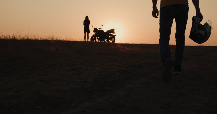 The silhouette of a man walking to the motorcycle where he is waiting for a young woman. Romantic travel concept