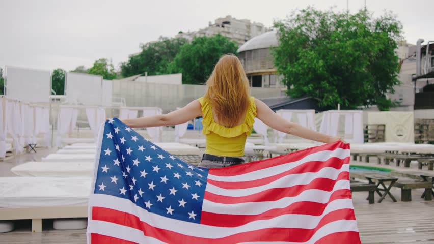 A beautiful young woman walks with an American flag along a beach club with swimming pools during a light summer rain. Girl rejoices celebrating US Independence Day
