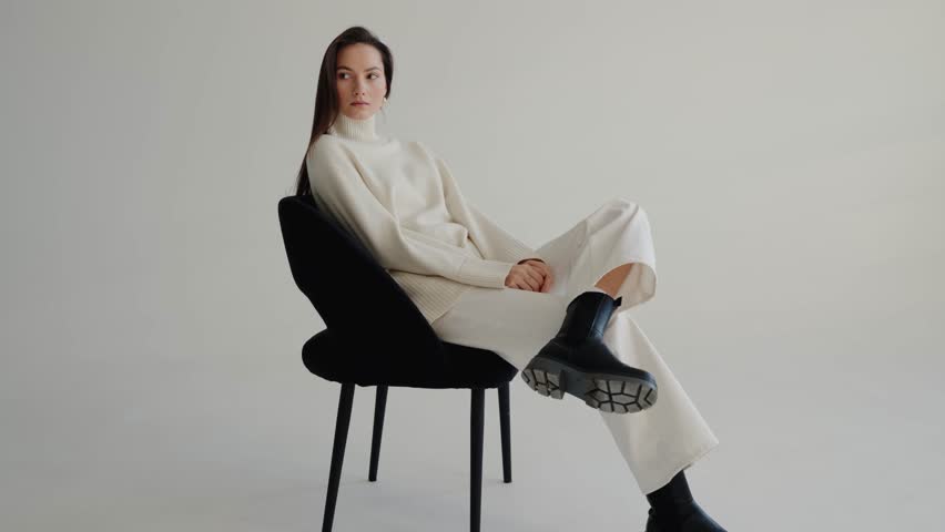 Beautiful young brunette woman with white sweater and pants sitting on a chair and posing at studio. Cute girl smiles on isolated white background. Fashion model with beautiful eyes.