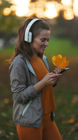 Hello autumn. happy stylish female in fitness clothes in the park with autumn leaf and headphones.