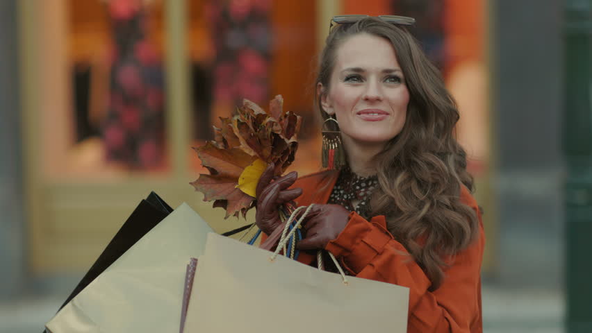 Hello october. smiling modern 40 years old woman in brown trench coat with shopping bags and autumn yellow leaves near shop in the city.