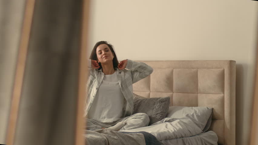 Caucasian European woman sitting on bed stretching good morning cheerful bedroom inside mirror pajamas cozy comfortable wellness carefree weekend relax resting awaken domestic holiday pleasure delight