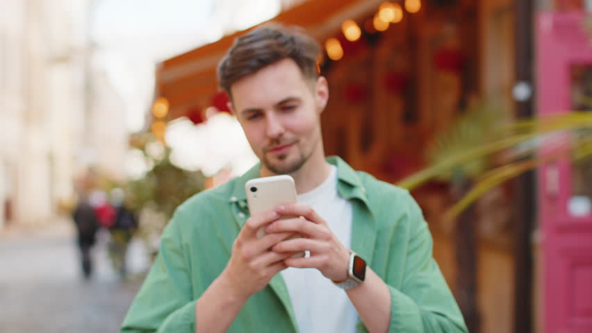 Smiling young man using smartphone typing text messages browsing internet, finishing work, looking at camera outdoors. Handsome guy on urban city street. Camera zooming in and person becomes in focus.