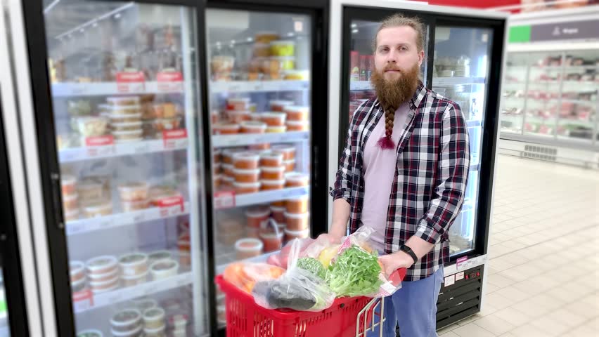 Man in a supermarket portrait. Man Young European Buyer Positive Bearded Long Haired, With Basket In Hands, Shopping Choosing Vegetables Organic In Supermarket.
