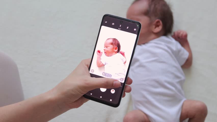 Mother shooting her newborn baby by smartphone lying on white sheet taking picture of baby with mobile phone captures the moments of the first days of his son's life