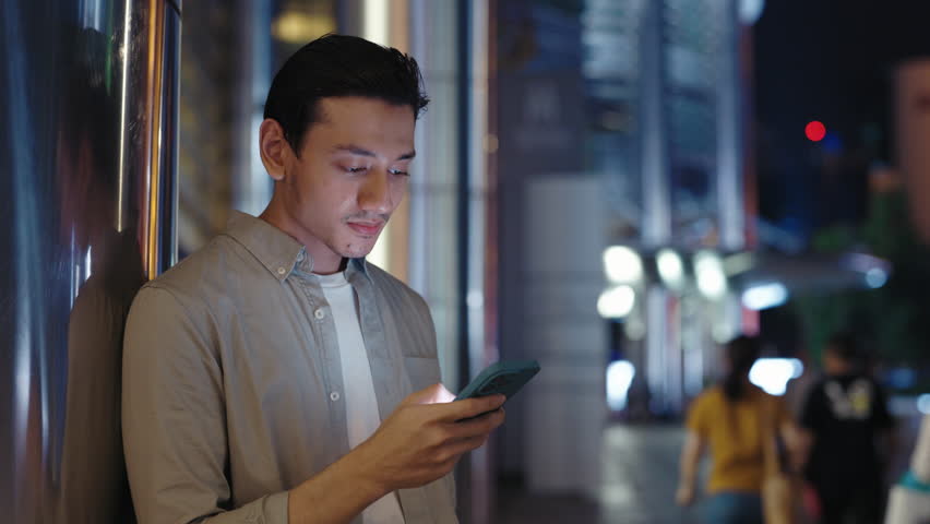 Handsome Asian Guy Scrolls Through News Feed on a Smartphone While Standing in Courtyard in City Center. Young Man Flipping Through Photos in Gallery Using Phone. Technology and Communication Concept
