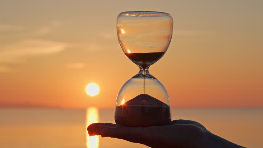 Hourglass concept time woman holding glass hourglass in palm on blurred background of golden sun disk sky in sunset rays in summer close-up. Golden sand pours quickly. Time concept sand. Relax