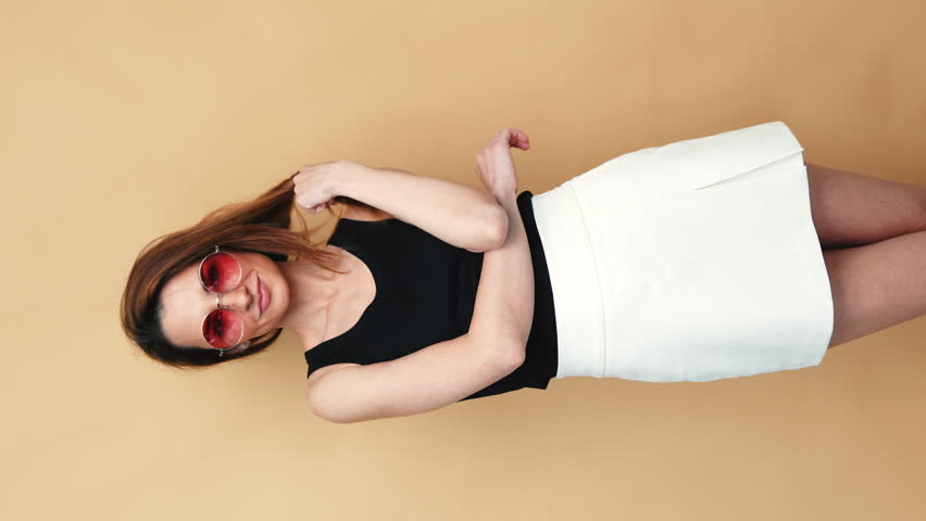 Woman in black t-shirt and white skirt. Sexy carefree stylish model with long hair. Smiling female posing in studio. Isolated. Looks delightful and cute. near beige wall. Vertical video