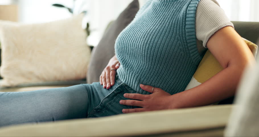 Woman, stomach and cramps pain in home for menstruation period, digestion or inflammation. Person, hands and bloated abdomen on sofa for uterus anatomy or stress anxiety for tummy ache, ulcer or hurt