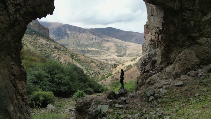 Woman stands under massive rock arch looking over hilly landscape