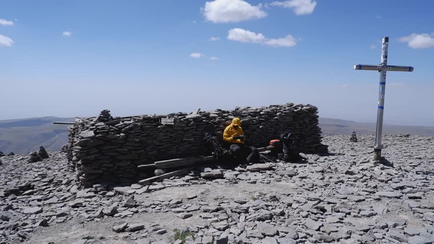 Climber rests by huge stone cairn near crucifix on mountain summit