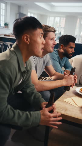 Vertical video of group of excited male friends at home in lounge watching sports on TV celebrating with beer and pizza together - shot in slow motion