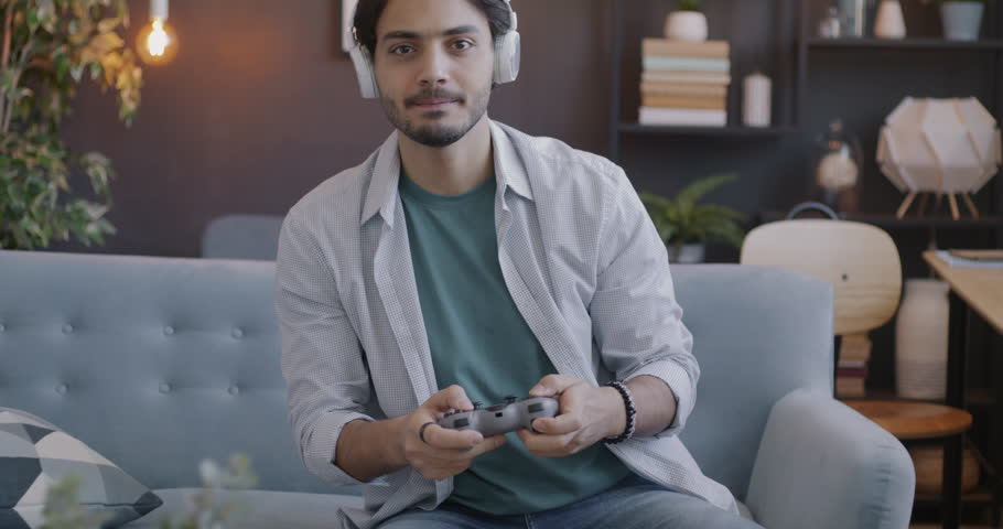 Portrait of Arab student playing video game enjoying leisure activity using headphones and controller at home in evening. People and gaming concept.