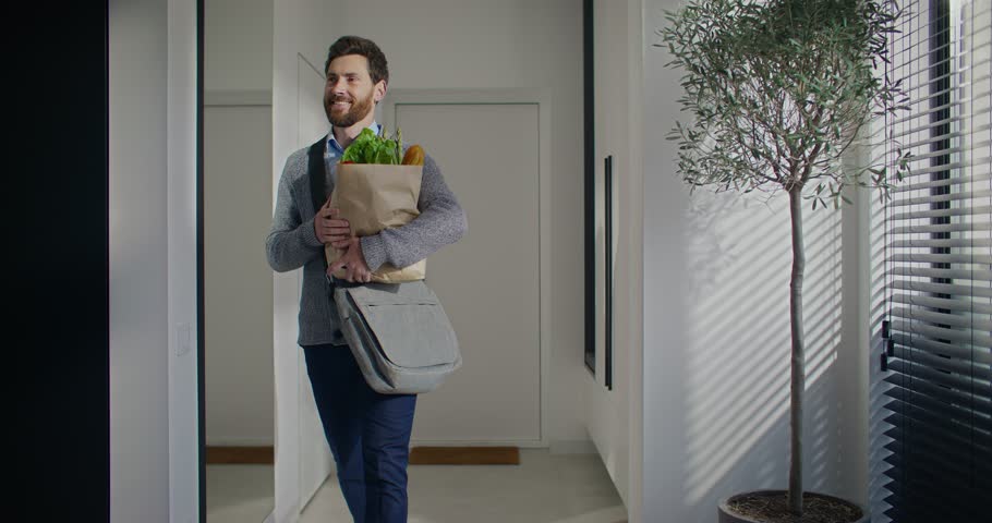 Charming Caucasian man entering his apartment. Dropping keys next to entrance at wardrobe. Walking through long foyer. Carrying paper bag full of groceries. Tired after hard working day.