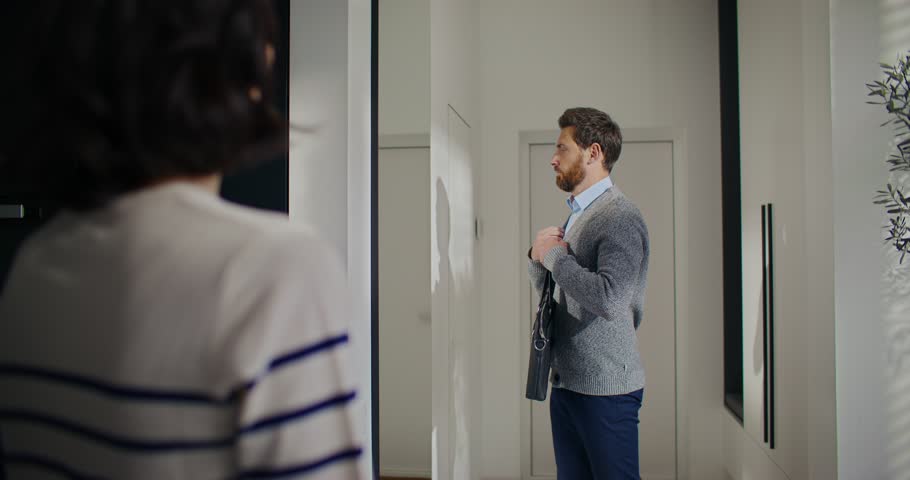 View from behind females shoulder. Wife looking at husband getting ready for work. Man checking outfit in front of big mirror. Holding briefcase while waving to beloved woman. Saying goodbye.