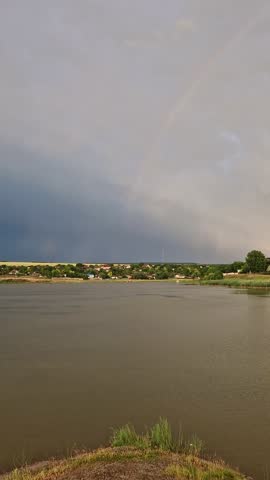 Storm clouds with lightning and a rainbow in the sky above the lake with a countryside view