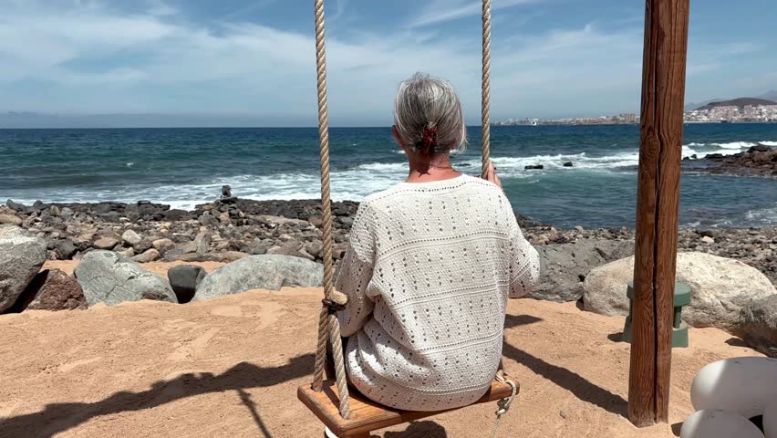 Happy senior woman sitting on swing on sea beach enjoying relaxed and carefree moments, retirement lifestyle for attractive senior lady