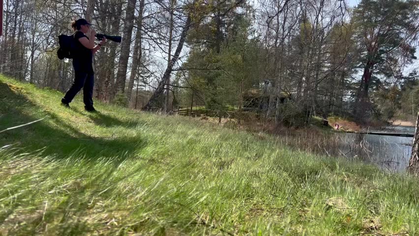 Low shot of a middle aged female photographer outside on a windy day lifting upp her camera fitted with a big sized lens which she points out to a body of water. Grass is blowing in the foreground. 