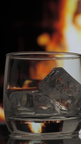 Pouring whiskey or scotch from a Bottle to a Glass over Fireplace background. Warmth and home comfort. vertical footage