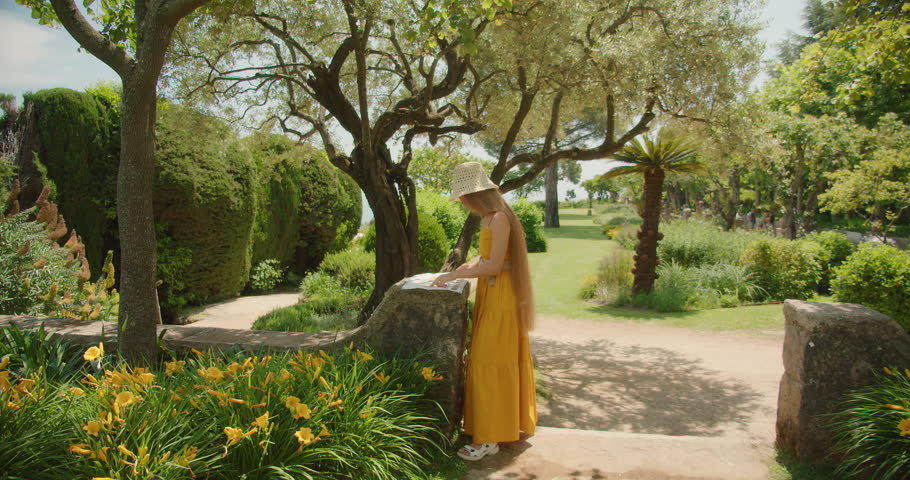 A woman in a yellow dress and hat studies a map on a stone pedestal in a lush garden, surrounded by trees and flowers.