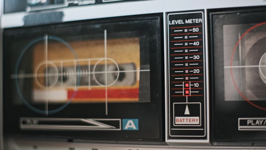Close-up of a vintage audio cassette tape player. The tape is playing and the level meter is bouncing. Red lights illuminate the scene, adding a retro vibe.