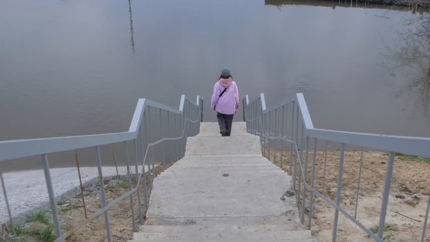 Woman walks down stairs to flooded river on spring day slow motion. Lady observes natural disaster consequences on city embankment upper view