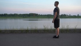 young man does a leg stretch while standing on the riverbank at the golden hour. handsome guy in sports clothes enjoys training outside in the summer.