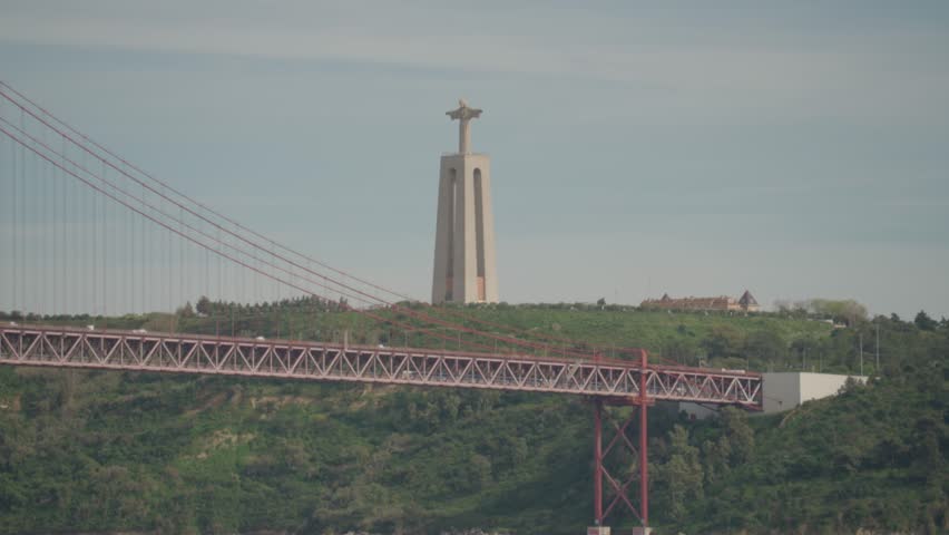 The majestic Santuário de Cristo Rei, or Christ the King monument, stands atop a hill with the iconic 25th of April Bridge in the foreground in Lisbon, Portugal.