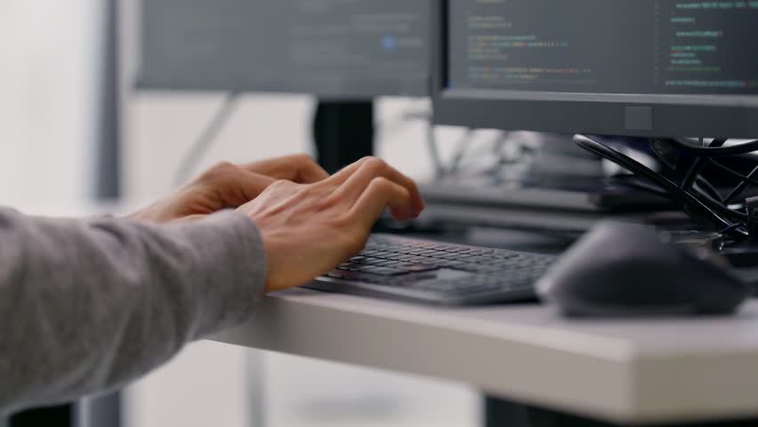 A close-up shot of a professional programmer’s hands typing code on a keyboard, with multiple monitors displaying lines of code in the background.