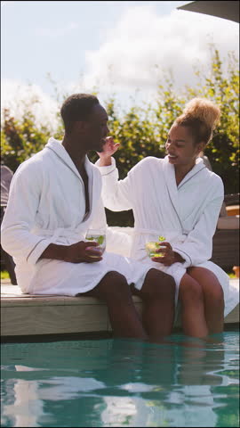 Vertical video portrait of couple wearing robes outdoors sitting and relaxing celebrating with drinks by swimming pool with feet in water on spa day - shot in slow motion