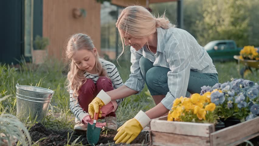 Pretty family working together during sunny day. Cute blond daughter with gardening tool digging hole for planting. Loving mother looking at work process and smiling. Teamwork.