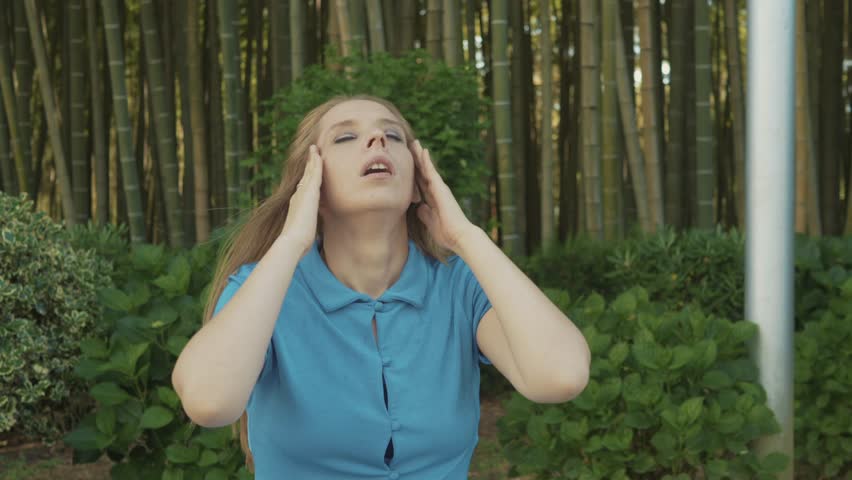 Young woman sitting in park, holding head with both hands, visibly experiencing anxiety and stress. Facial expression and body language indicate mental health struggles