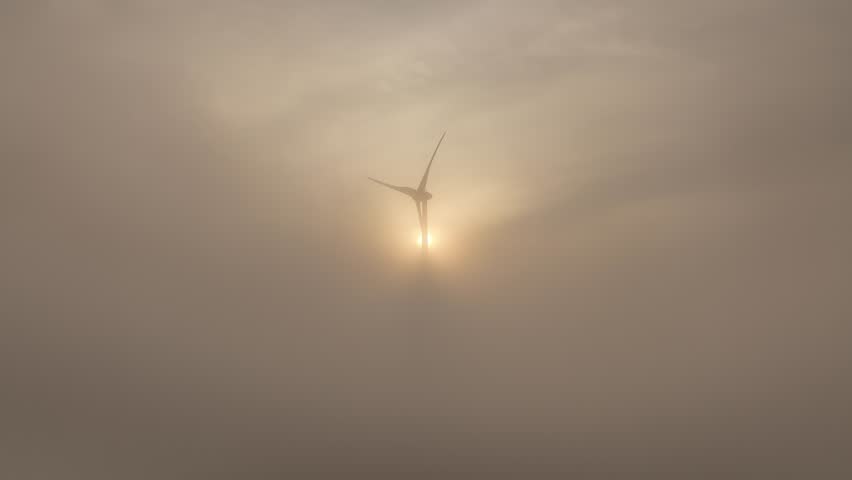 A drone footage of a wind turbine covered with dense fog with the sunrise scene in the background