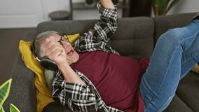 Joyful senior man with gray hair wearing headphones and glasses, laughing while lying on a sofa in a cozy living room at home. - Powered by Shutterstock - Get 15% off with code: PIKWIZARD15